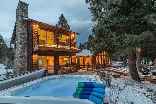 Modern log cabin with stone chimney, illuminated windows, hot tub in foreground, surrounded by evergreen trees and snow.