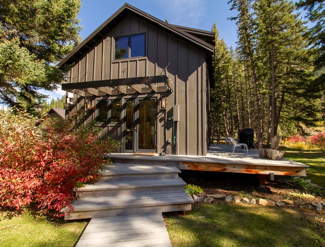 Modern black and tan cabin with elevated deck surrounded by tall pine trees and red autumn foliage